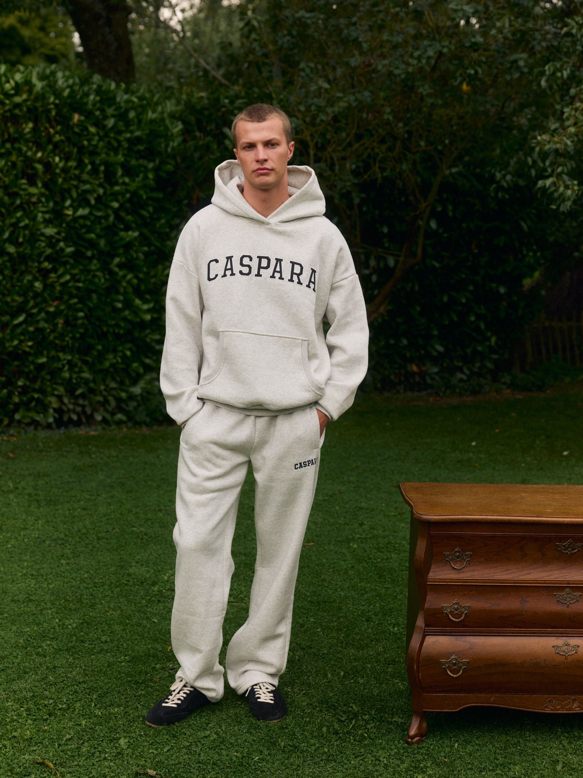 Man wearing a grey melange hoodie and straight-leg sweatpants standing on grass next to a wooden dresser.